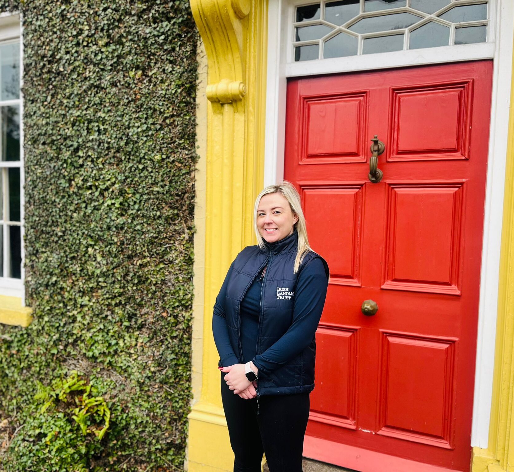 Photo of Stacey Reilly, a blonde lady in Irish Landmark Trust branded gilet, standing outside red door of Tullymurry House