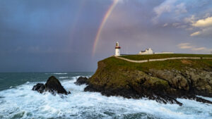 Wide shot of Galley Head Lighthouse at Duneady Head in Cork, a rainbow is shining over the lighthouse, behind it a dramatic sky and below the sea crashes against the headland