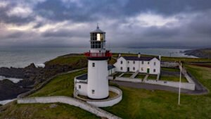 Galley Head Lighthouse on a moody day with the sun peeking through purple cloudy skies, the lighthouse is dramatic in the foreground with the two houses for rent facing it.